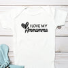 A white short-sleeve baby bodysuit laid flat on a white wood plank background, printed with the phrase 'I LOVE MY Ammamma' and a heart graphic in black. A light blue plush elephant toy with black-and-white stripes is placed beside it.
