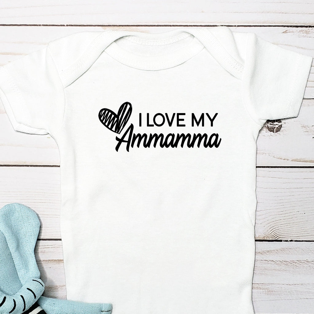 A white short-sleeve baby bodysuit laid flat on a white wood plank background, printed with the phrase 'I LOVE MY Ammamma' and a heart graphic in black. A light blue plush elephant toy with black-and-white stripes is placed beside it.