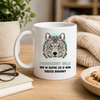 A white ceramic mug with a printed gray wolf illustration and Polish text, placed on a wooden table in a cozy indoor setting with books, glasses, cookies, and a knit blanket in the background.