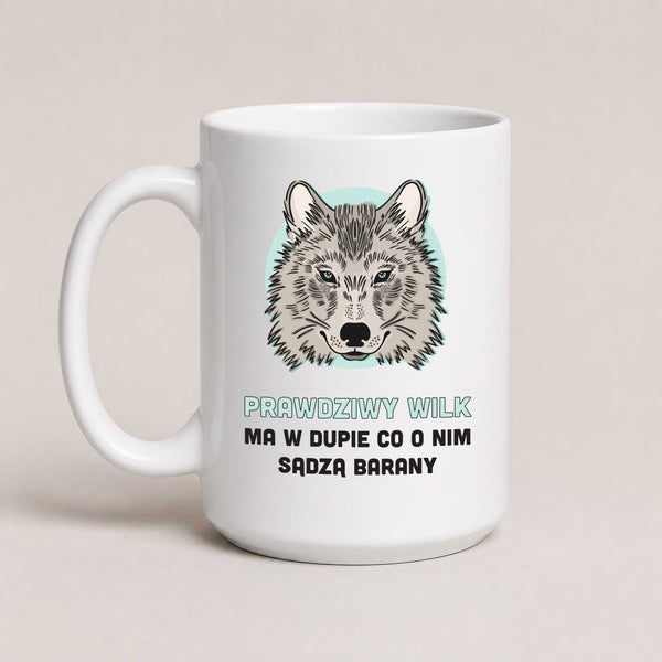 A white ceramic mug with a printed graphic of a wolf's face on a light blue circular background, above Polish text reading 'PRAWDZIWY WILK MA W DUPIE CO O NIM SĄDZA BARANY'. The mug is photographed against a plain, warm off-white background.